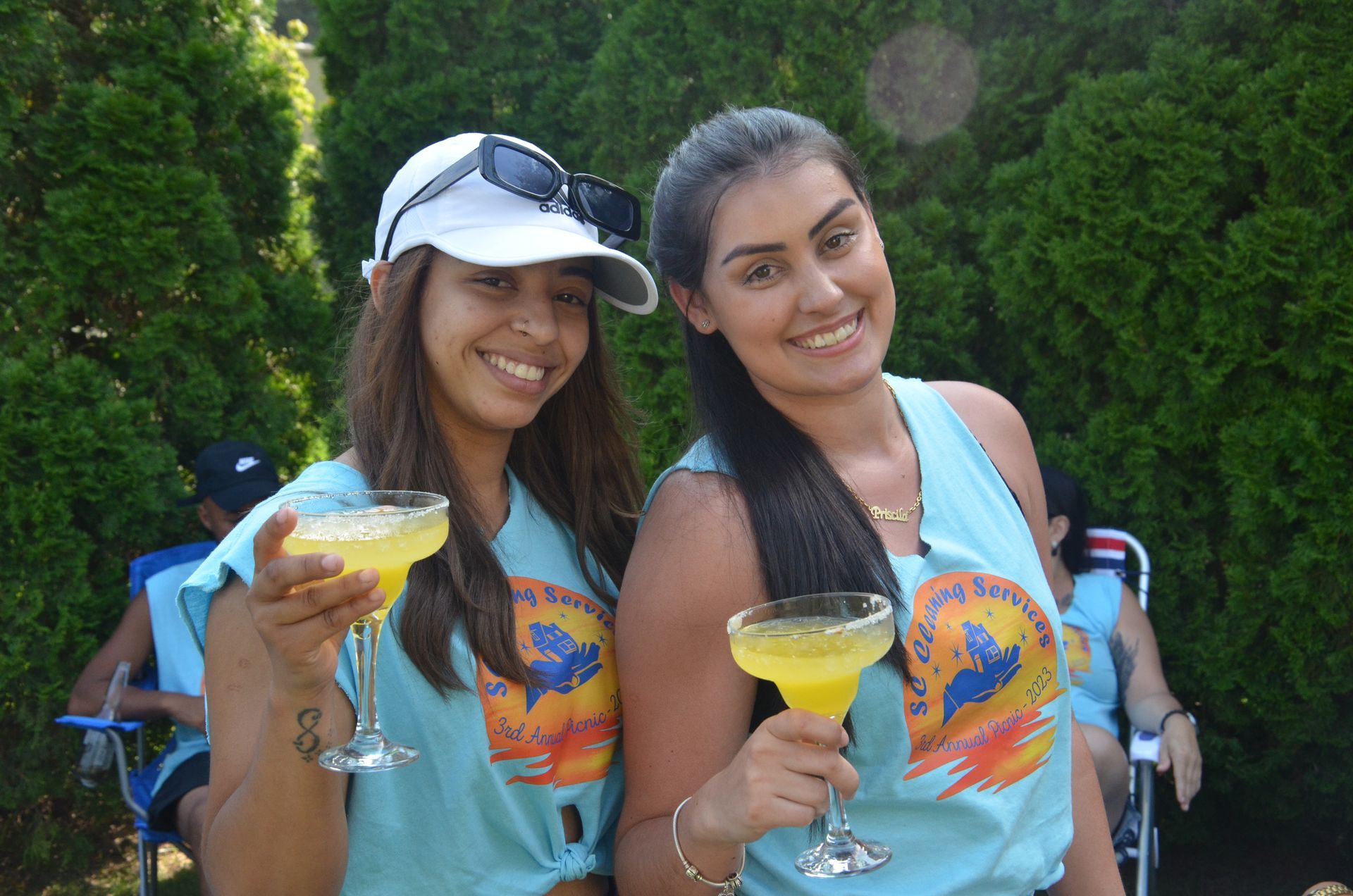 Two smiling women holding margaritas, wearing matching blue shirts outdoors.