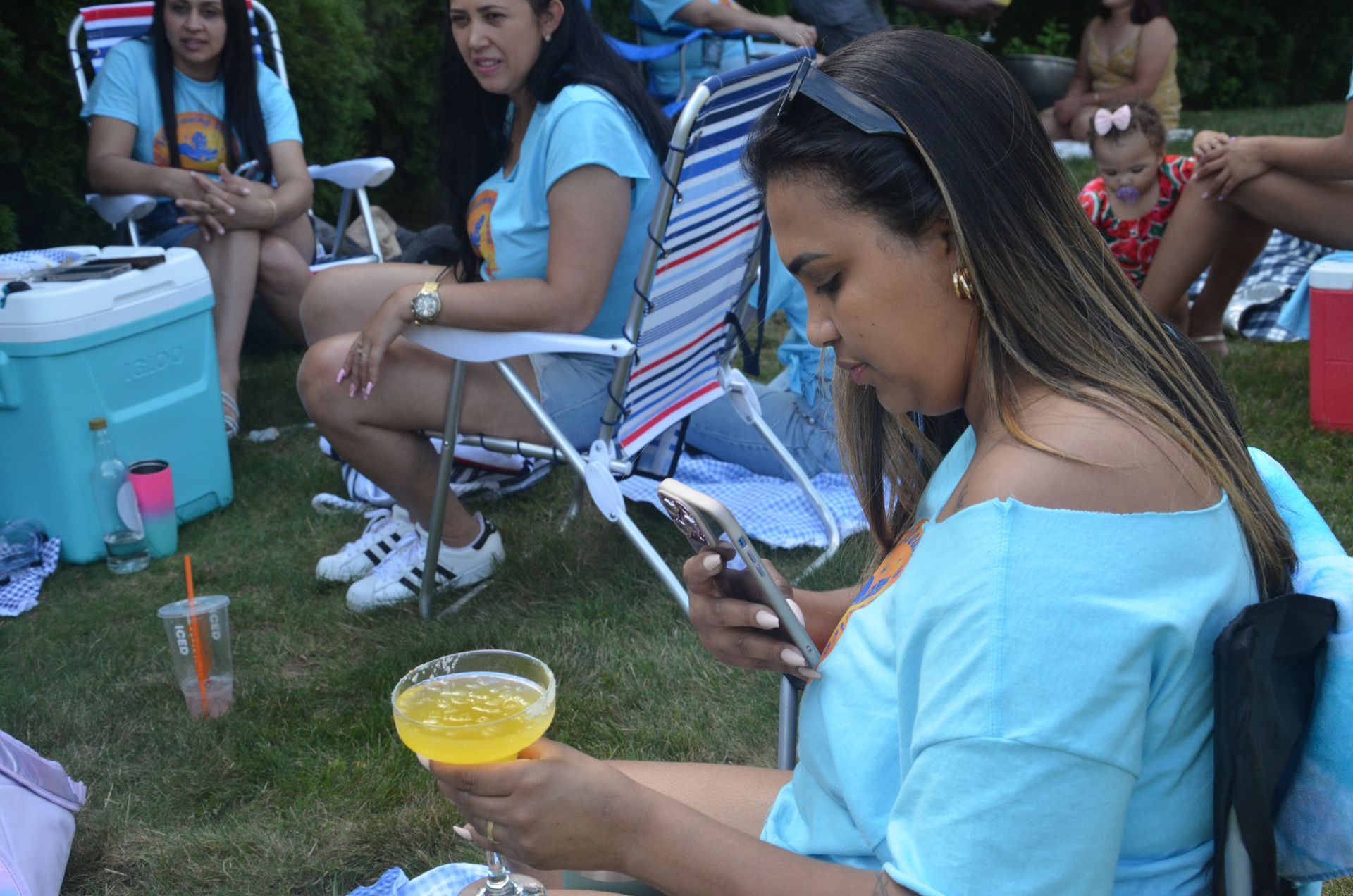 Woman in blue shirt, holding a drink and phone, in a grassy outdoor gathering. People in chairs, picnic.