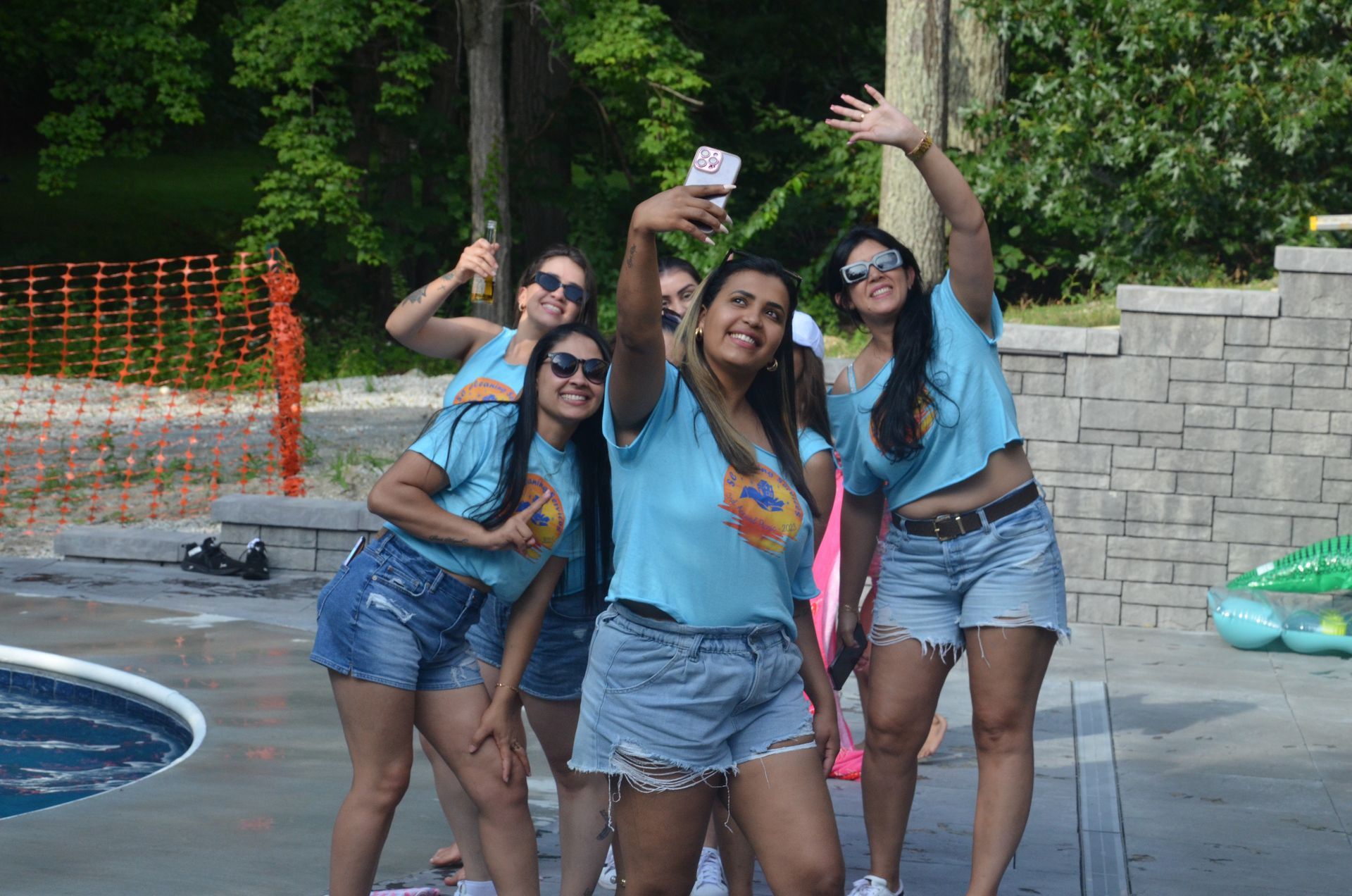 Five women in matching blue shirts and jean shorts take a selfie near a pool, waving and smiling.
