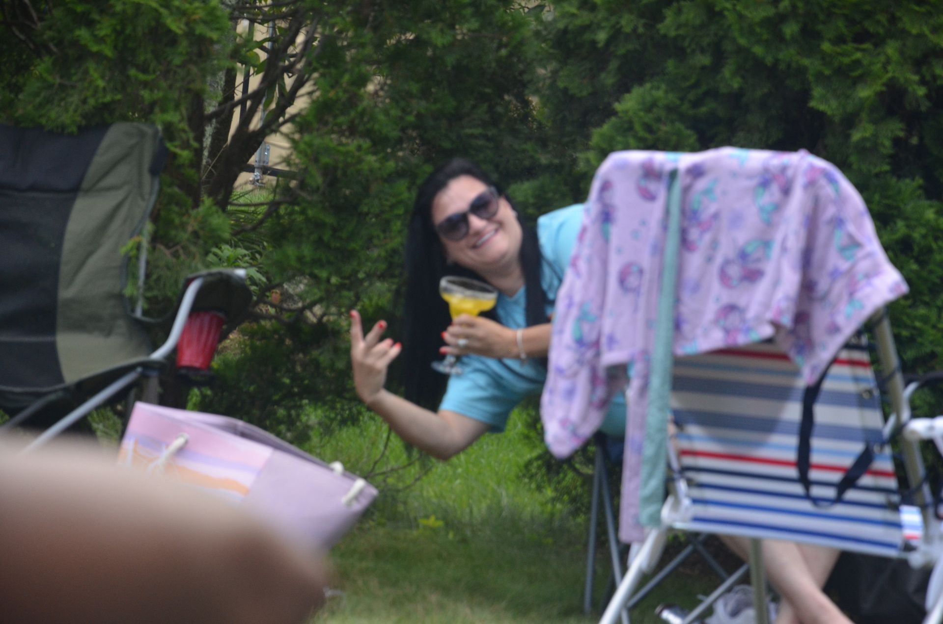 Woman in sunglasses holds a drink, making a peace sign, outdoors with lawn chairs.