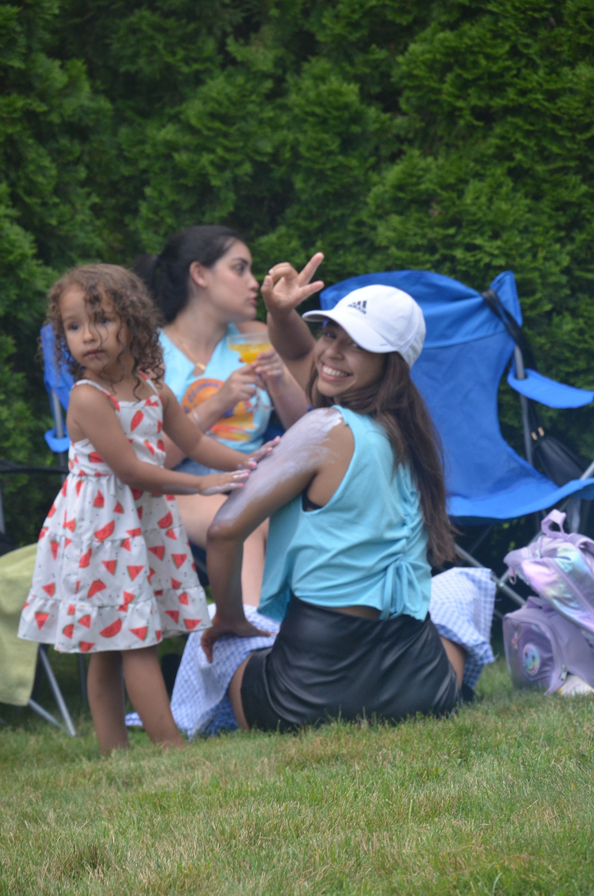 Woman applying sunscreen, child nearby, another woman in background, outdoor setting.