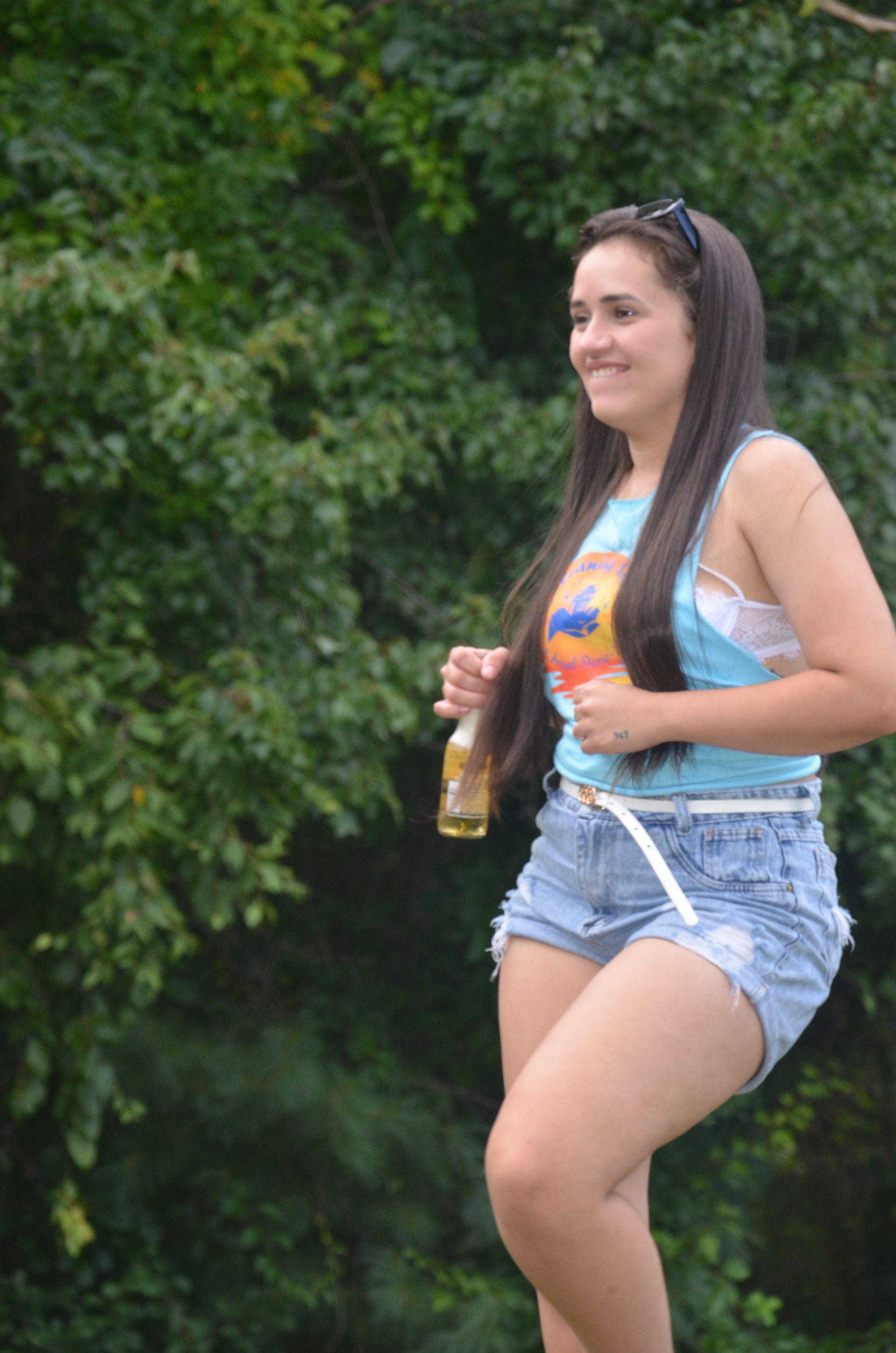 Woman in jean shorts and tank top holds a beer bottle, smiling against a green backdrop of trees.