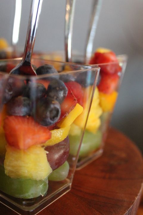 Fruit salad in clear cups with spoons, on a wooden tray; layered with grapes, pineapple, berries, and more.