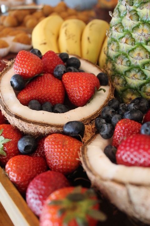 Close-up of fresh strawberries and blueberries in halved coconut shells, with bananas and a pineapple.