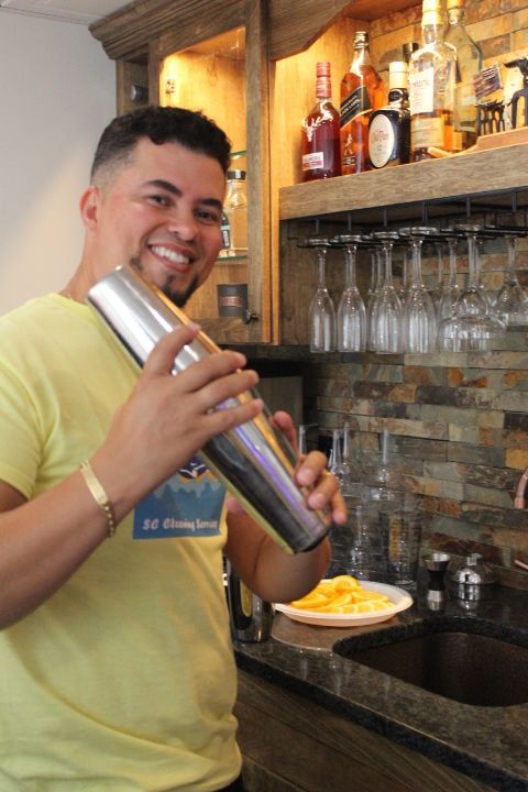 Man in yellow shirt shaking a cocktail shaker in a bar, smiling.