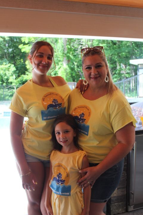 Three women in yellow shirts smile in front of a building with trees in the background.