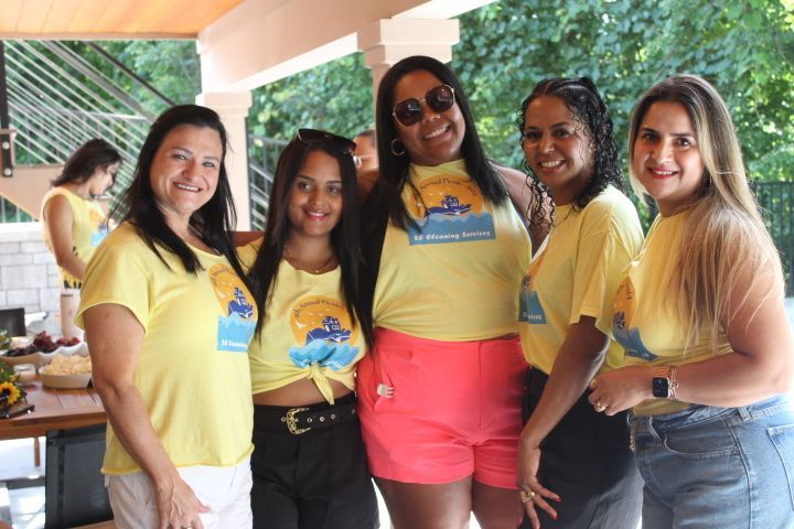 Five smiling women in yellow shirts pose outdoors. One wears pink shorts, others jeans or black pants.