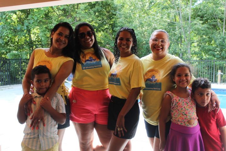 Group of six women and two children smiling for the camera, wearing yellow shirts, standing near a pool.