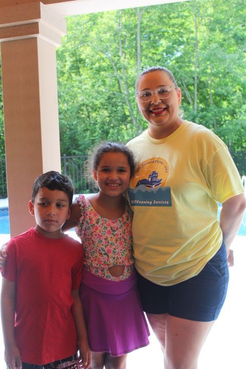 Woman, two children pose outdoors by a pool; woman wears yellow shirt, kids wear swimsuits.