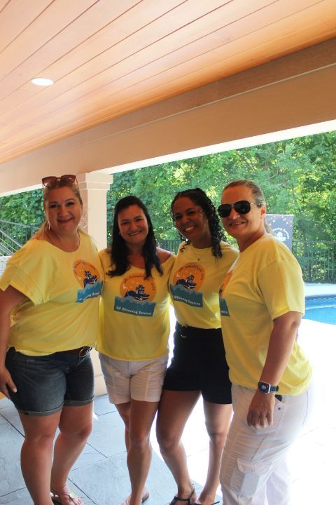 Four women in yellow shirts, smiling, stand on a patio near a pool.