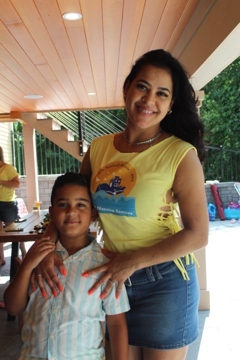 Woman and boy smiling outdoors, woman's hand on boy's shoulder. Woman in yellow shirt and denim skirt, boy in button-up.