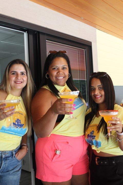 Three women in yellow shirts and shorts toasting drinks outdoors.