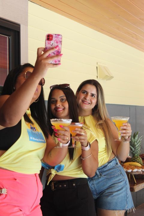 Three women taking a selfie, holding cocktails, wearing yellow tops, and posing indoors.