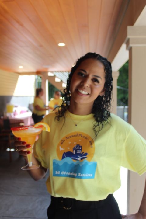 Woman in yellow shirt holding a margarita, smiling. Outdoors, event setting.