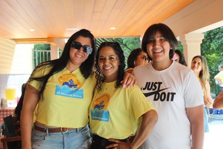 Three smiling women pose outdoors; two wear yellow shirts, one wears a 