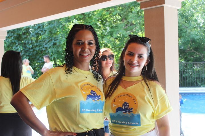 Two smiling women in yellow shirts pose outdoors. Trees and a swimming pool are in the background.