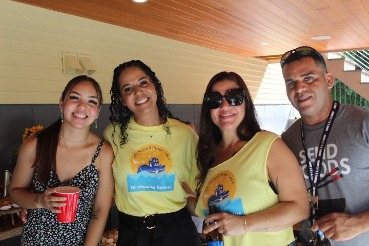 Four people smiling, posing for a photo. Outdoors, two wearing matching yellow shirts with a blue logo.