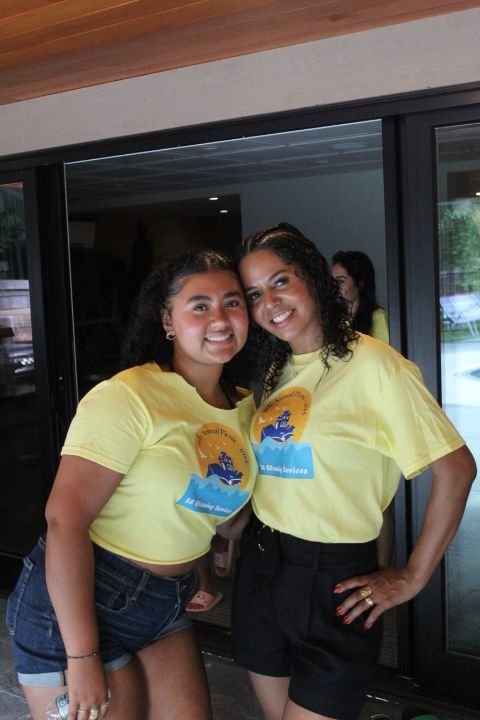 Two women in yellow t-shirts, dark shorts, smiling. Outdoors, near a sliding glass door.