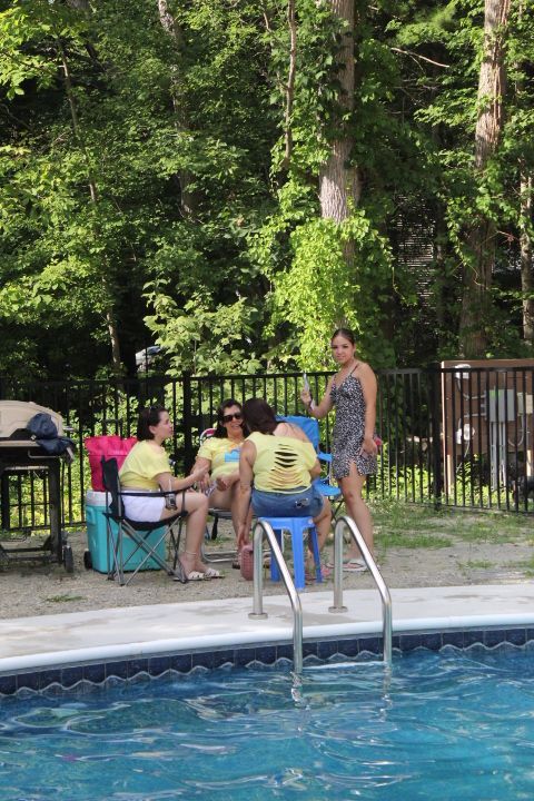 People near a pool, some wearing yellow shirts, chatting. Lush green trees in the background.