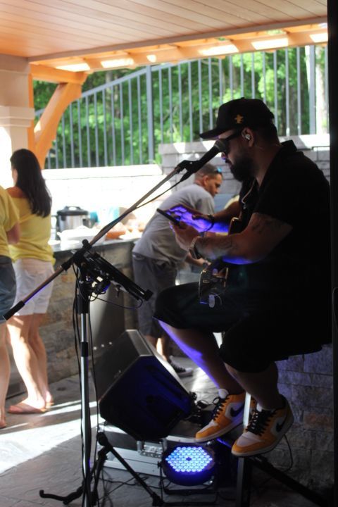 Man playing guitar and singing, outdoors. He wears a black hat, shirt, and shorts. Yellow sneakers.