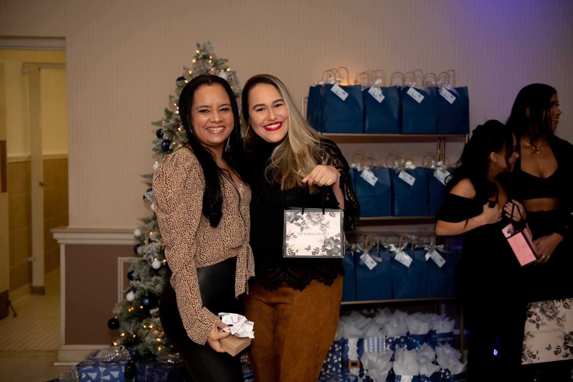 Two women smiling, posing with gifts near a Christmas tree and gift bags.