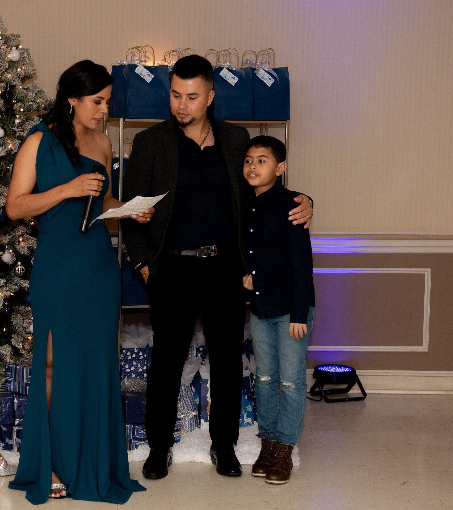 Woman in blue dress speaks, man and boy stand by her at event with Christmas decorations.