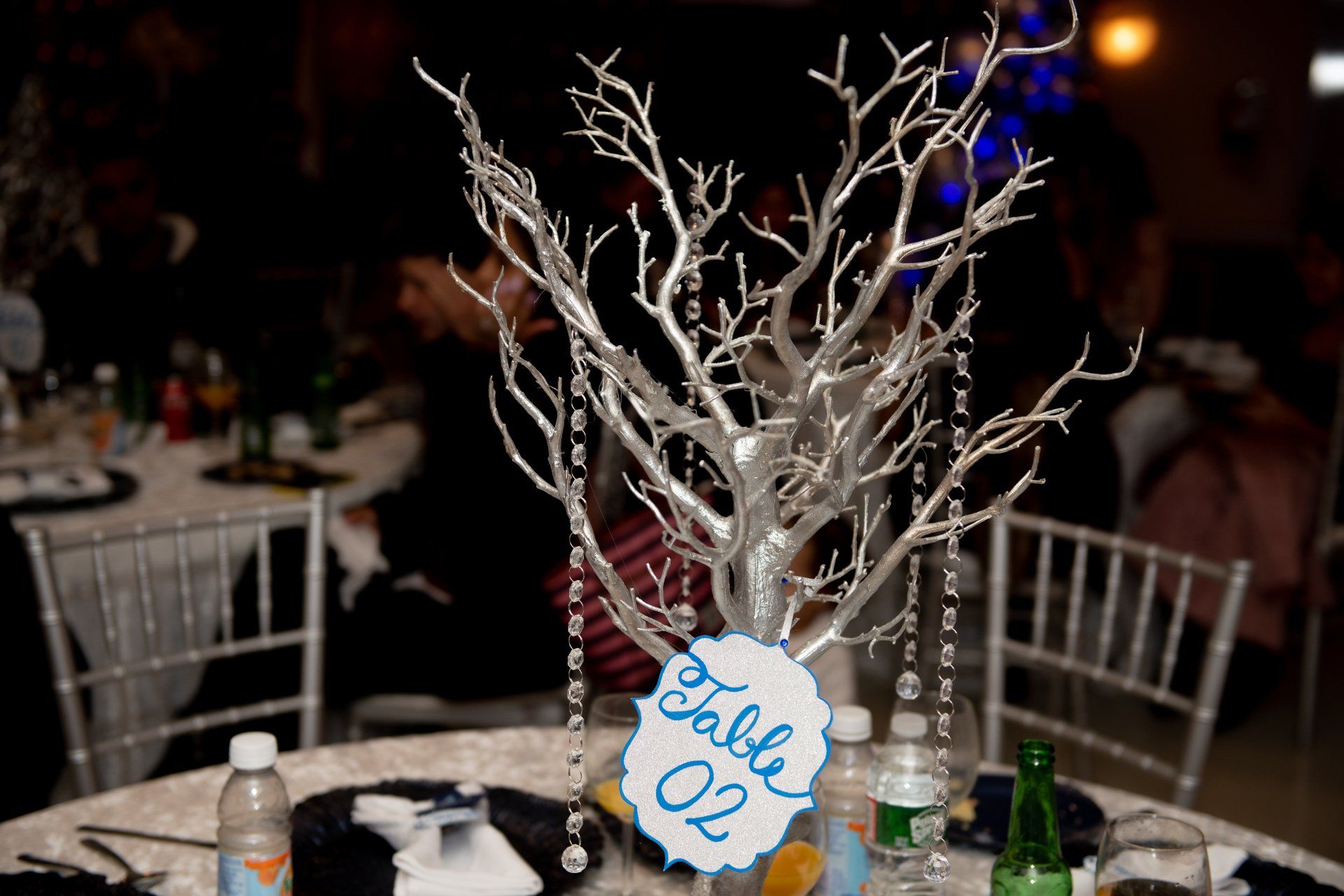 Silver tree centerpiece with hanging crystals; table 02 sign.  Dining table in a dim event setting.