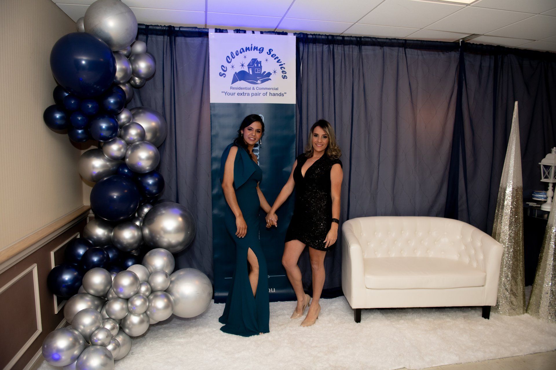 Two women pose by a balloon arch and sofa at an event; one in a blue dress, the other in black.