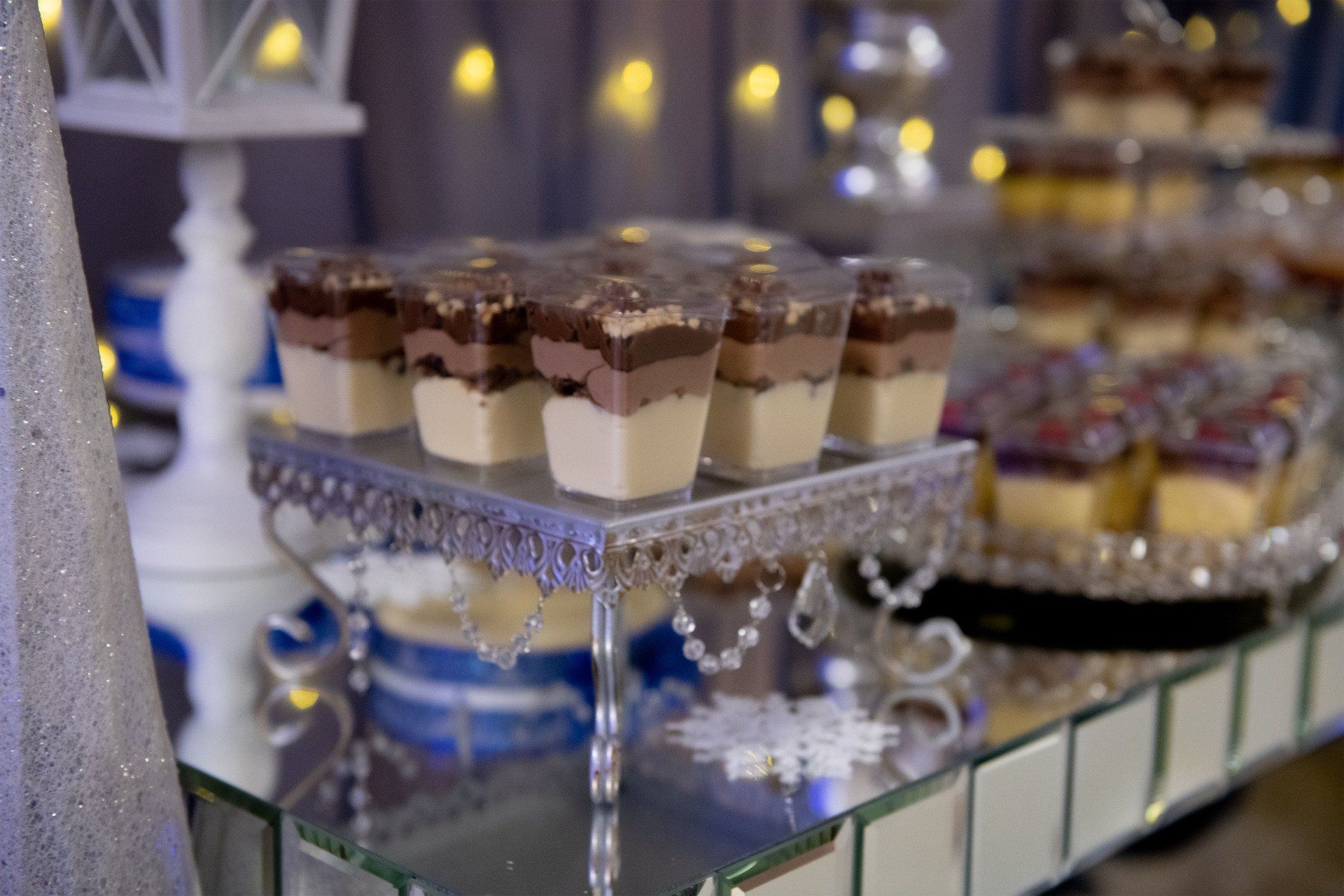 Dessert table with layered chocolate parfaits in clear cups on a silver stand; blurred string lights in the background.