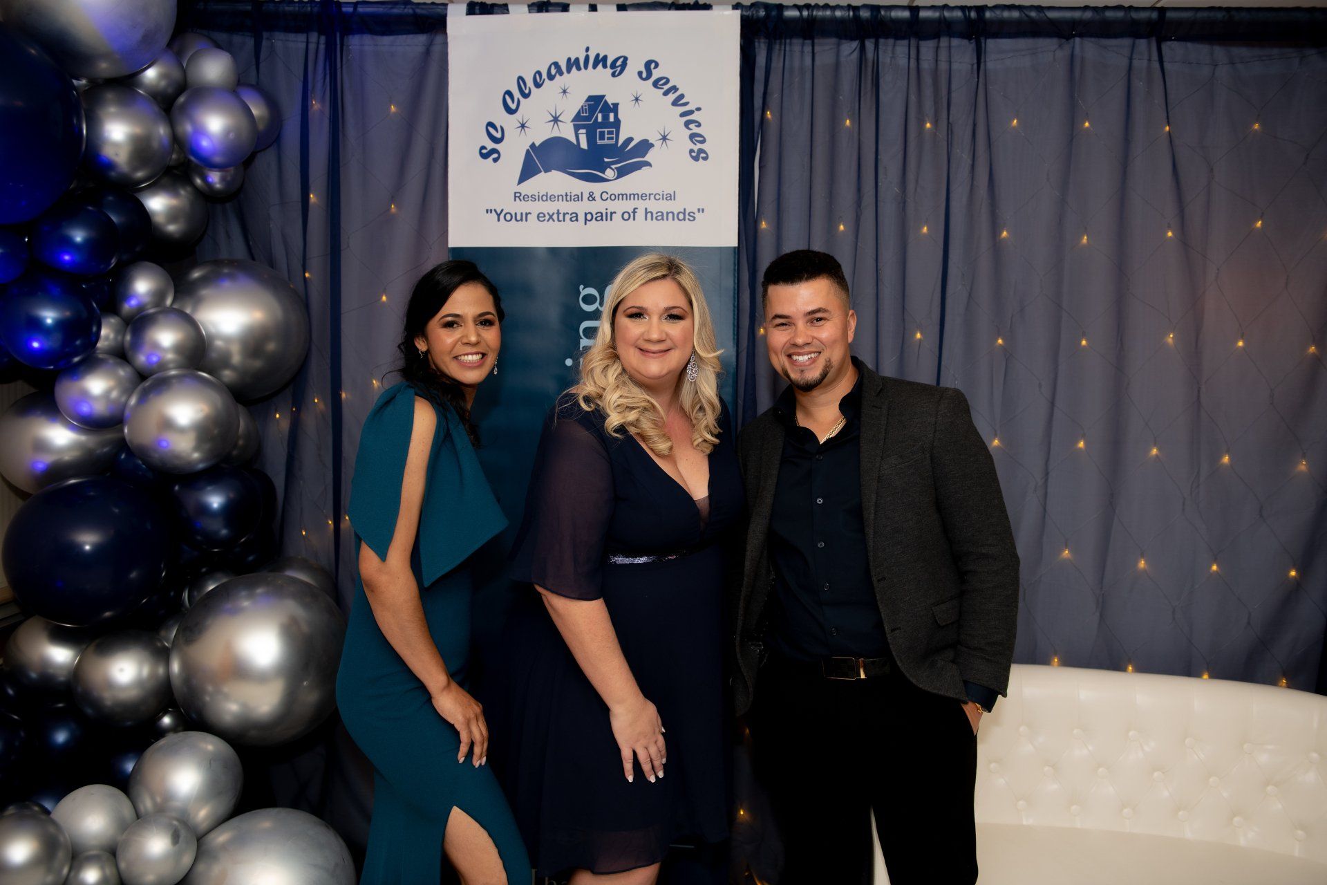 Three people pose in front of balloons and a banner. The women wear formal dresses. The man has a jacket.