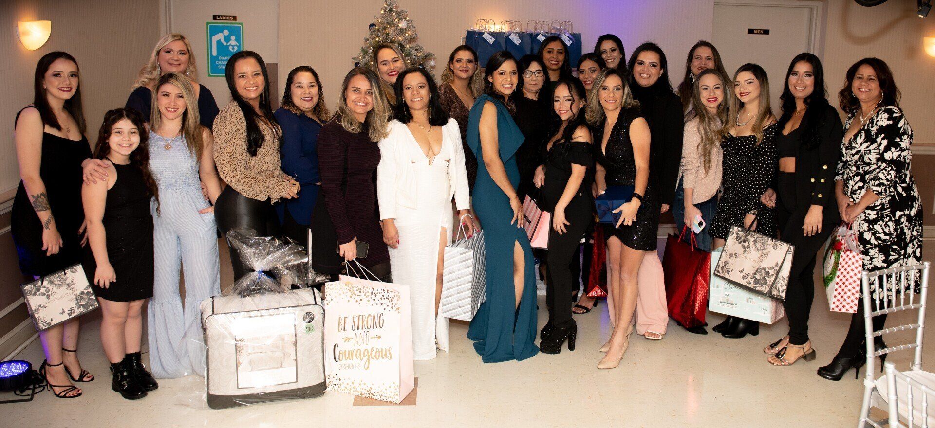 A large group of elegantly dressed women pose for a photo in a decorated room, some holding gift bags.