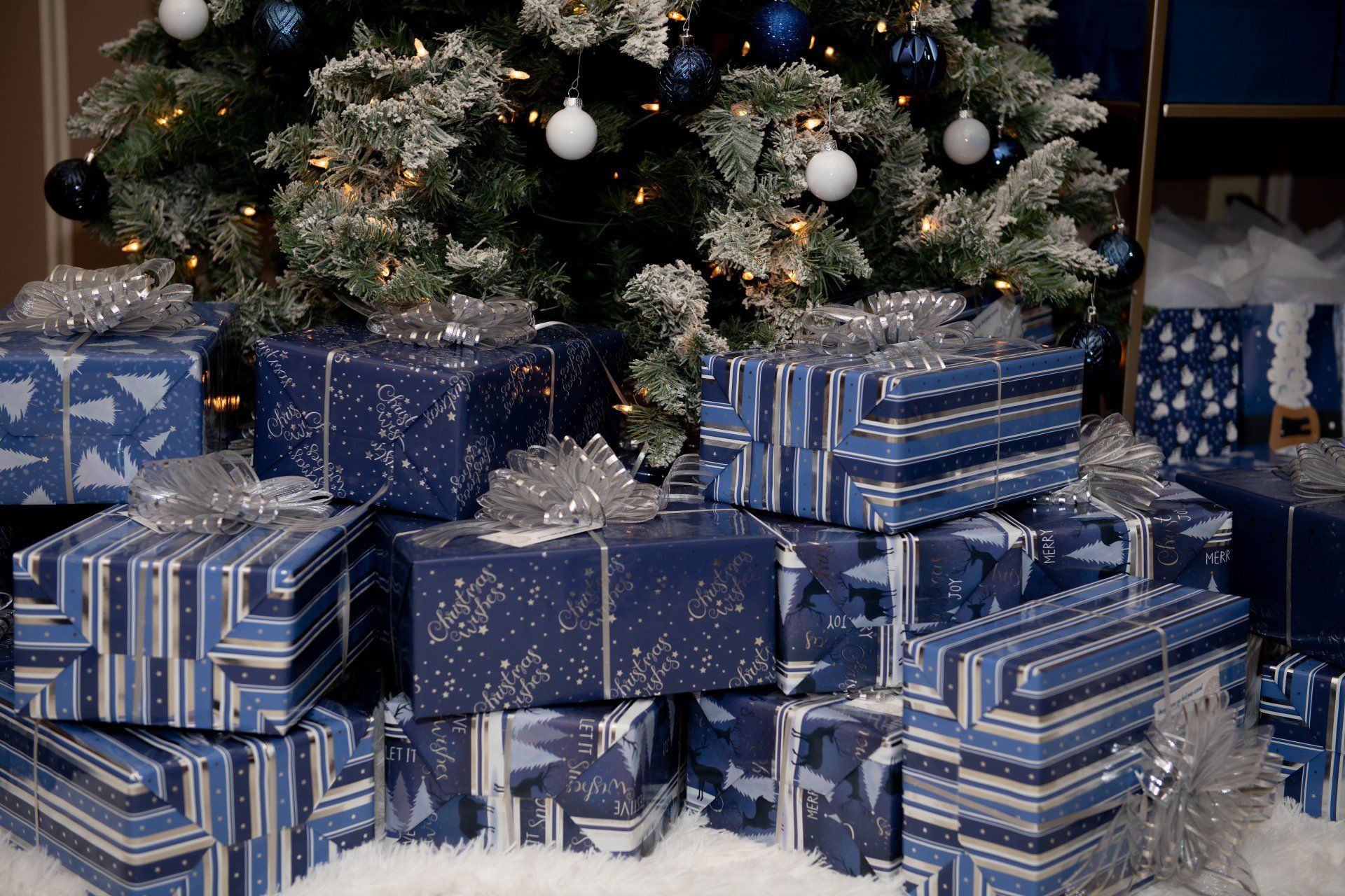 Blue and silver Christmas gifts under a snowy, decorated tree, with silver bows.