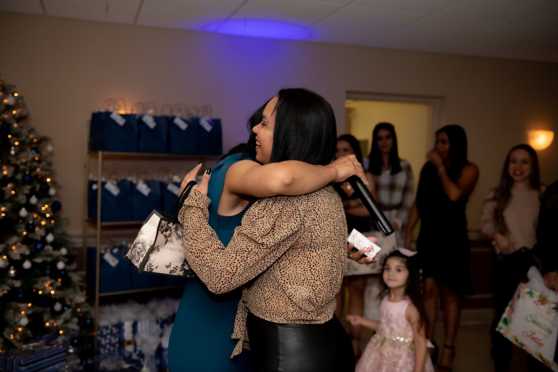 Two women embrace indoors. A microphone, a Christmas tree, and people are visible.