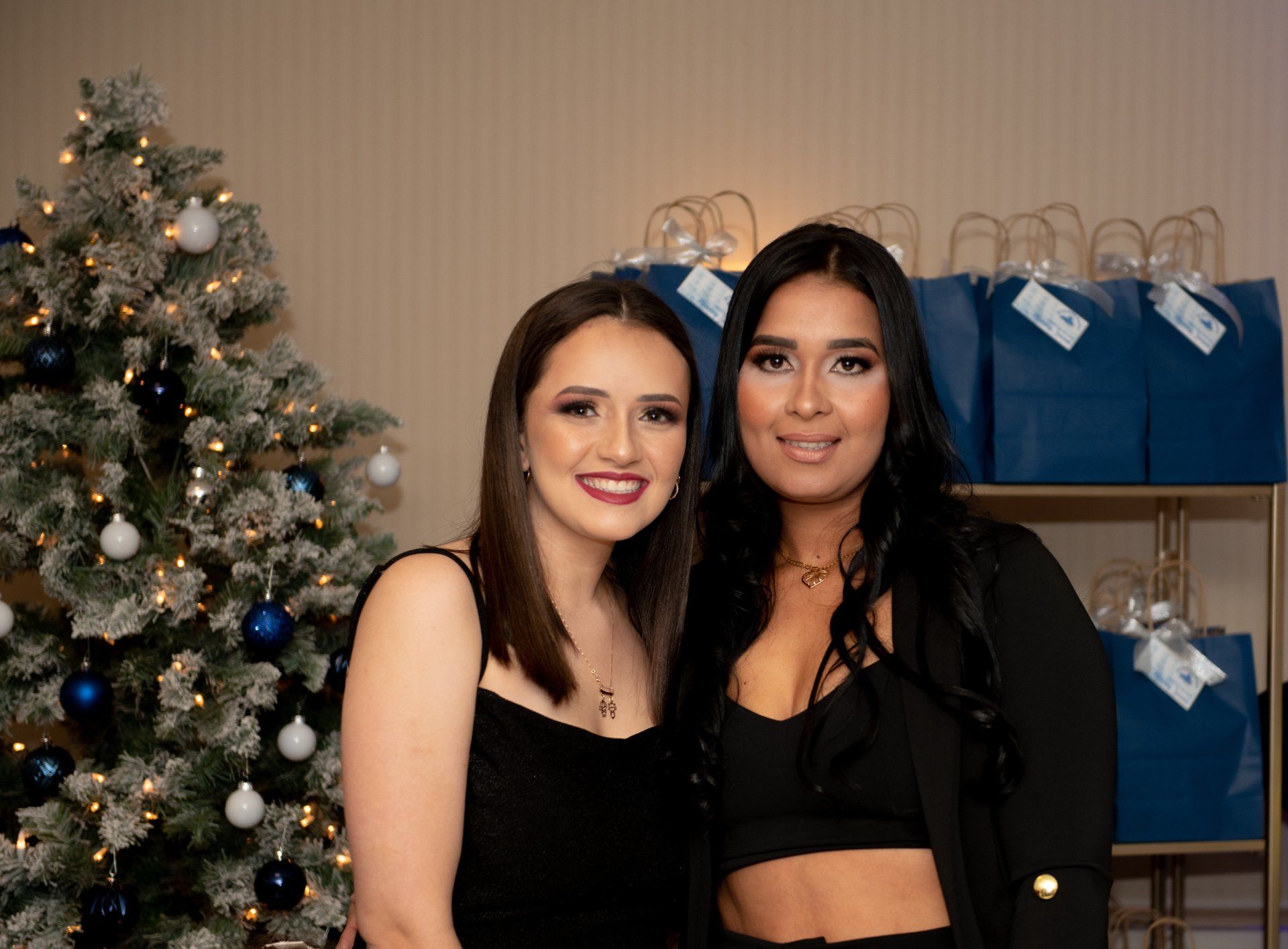 Two women smile in front of a Christmas tree and blue gift bags. One wears a black dress, the other a black top and blazer.
