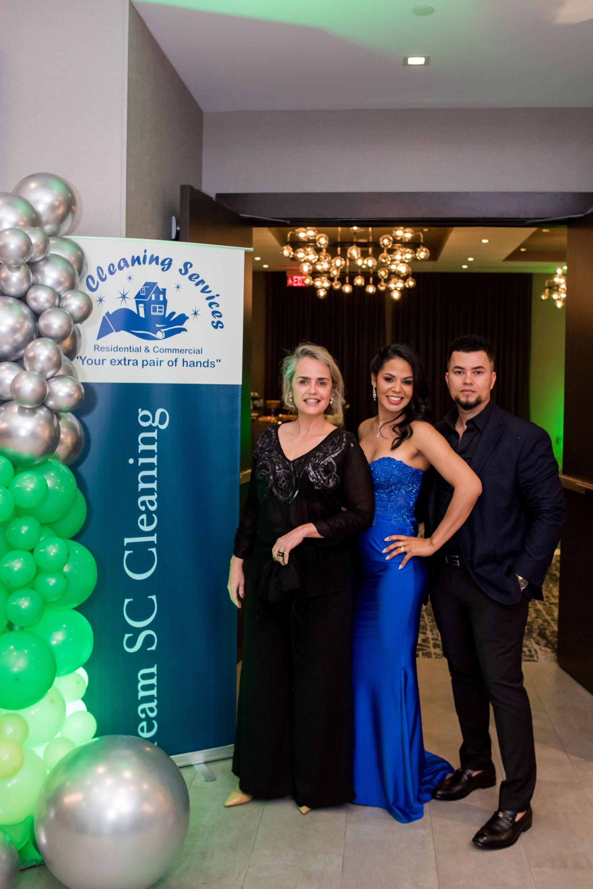 Three people pose by a banner with balloons. Woman in black dress, woman in blue gown, and man in navy shirt.