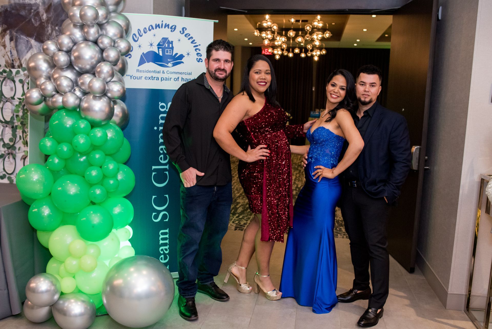 Four people pose in front of a banner, balloons. Two women wear dresses, two men wear shirts, a doorway in background.