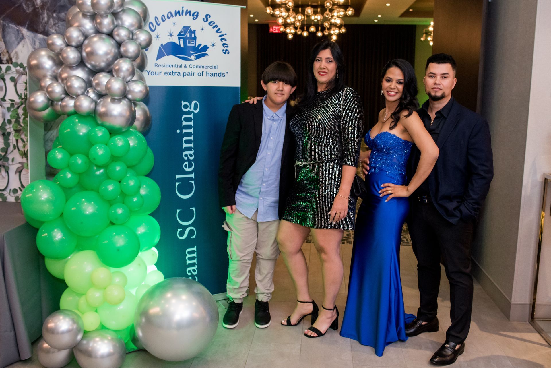 Four people pose at a party with balloons. One in a blue gown, others in party attire.