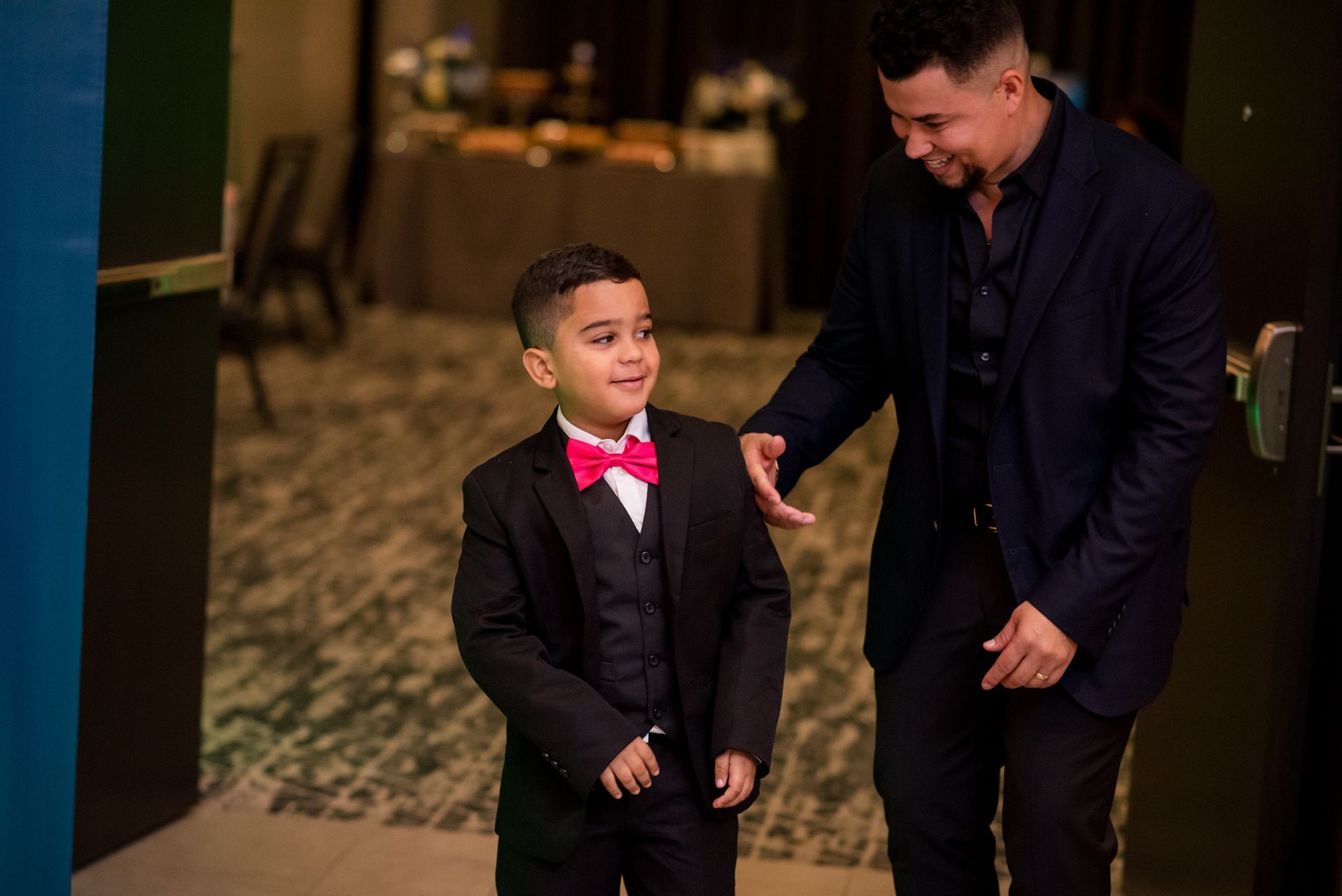 Father and son in formal wear. The boy smiles, wearing a tuxedo and pink bow tie.