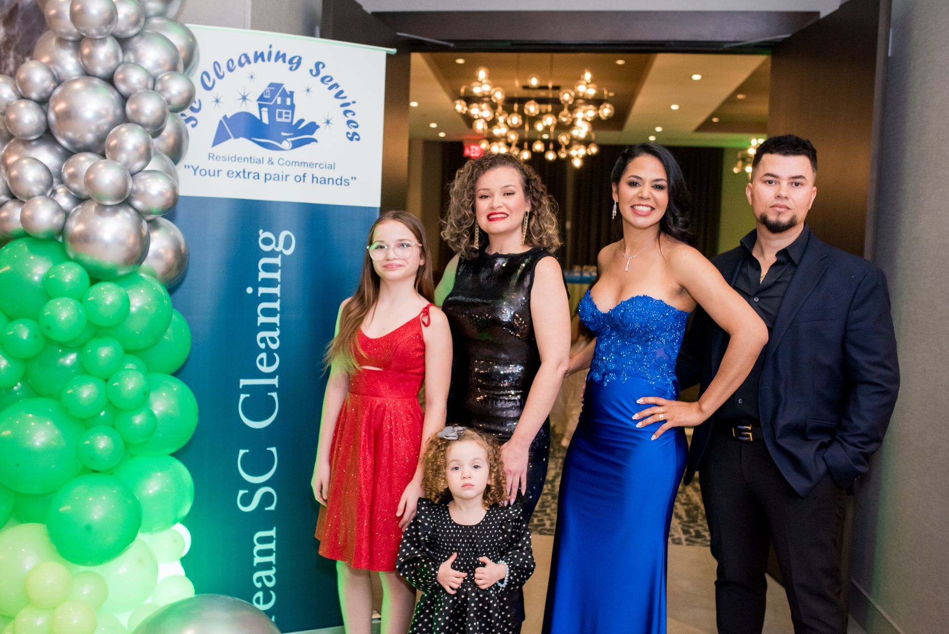 A group of five people posing in front of a banner, wearing formal attire. Green and silver balloons on left.