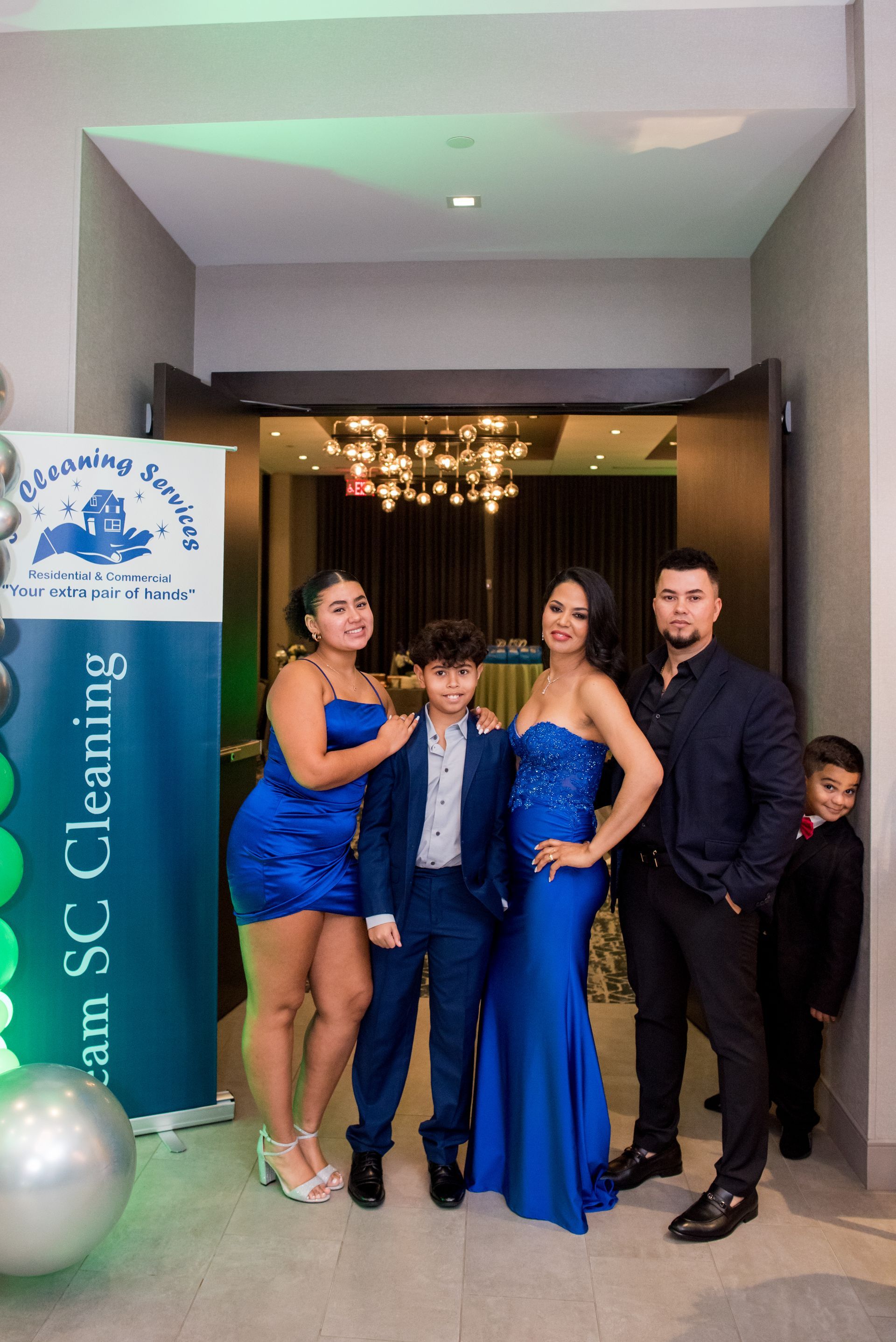 Family poses inside, near a banner; woman in blue gown, others in formal attire.