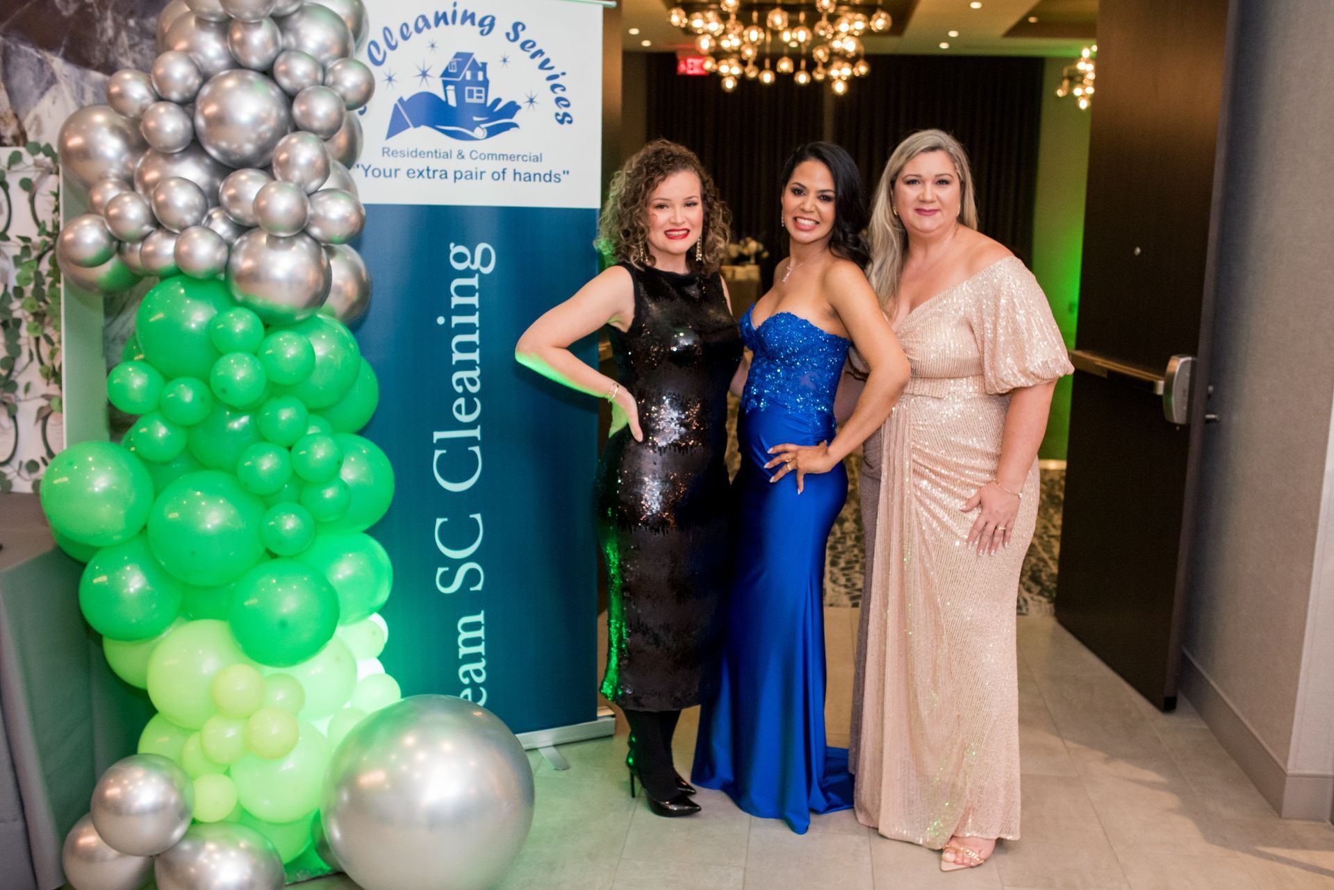 Three women in evening gowns pose next to a banner with balloons.