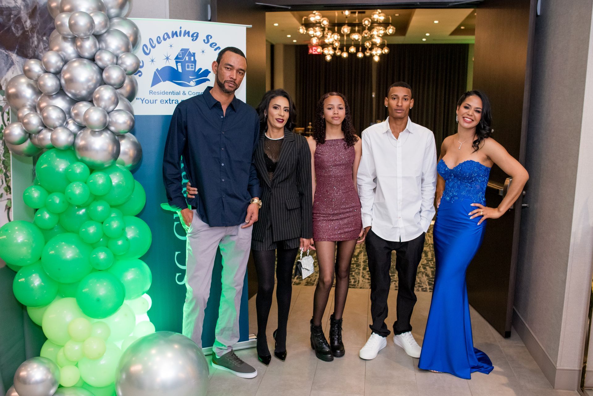 Five people pose in front of a banner with balloons. Formal wear, smiles. Indoors, party setting.