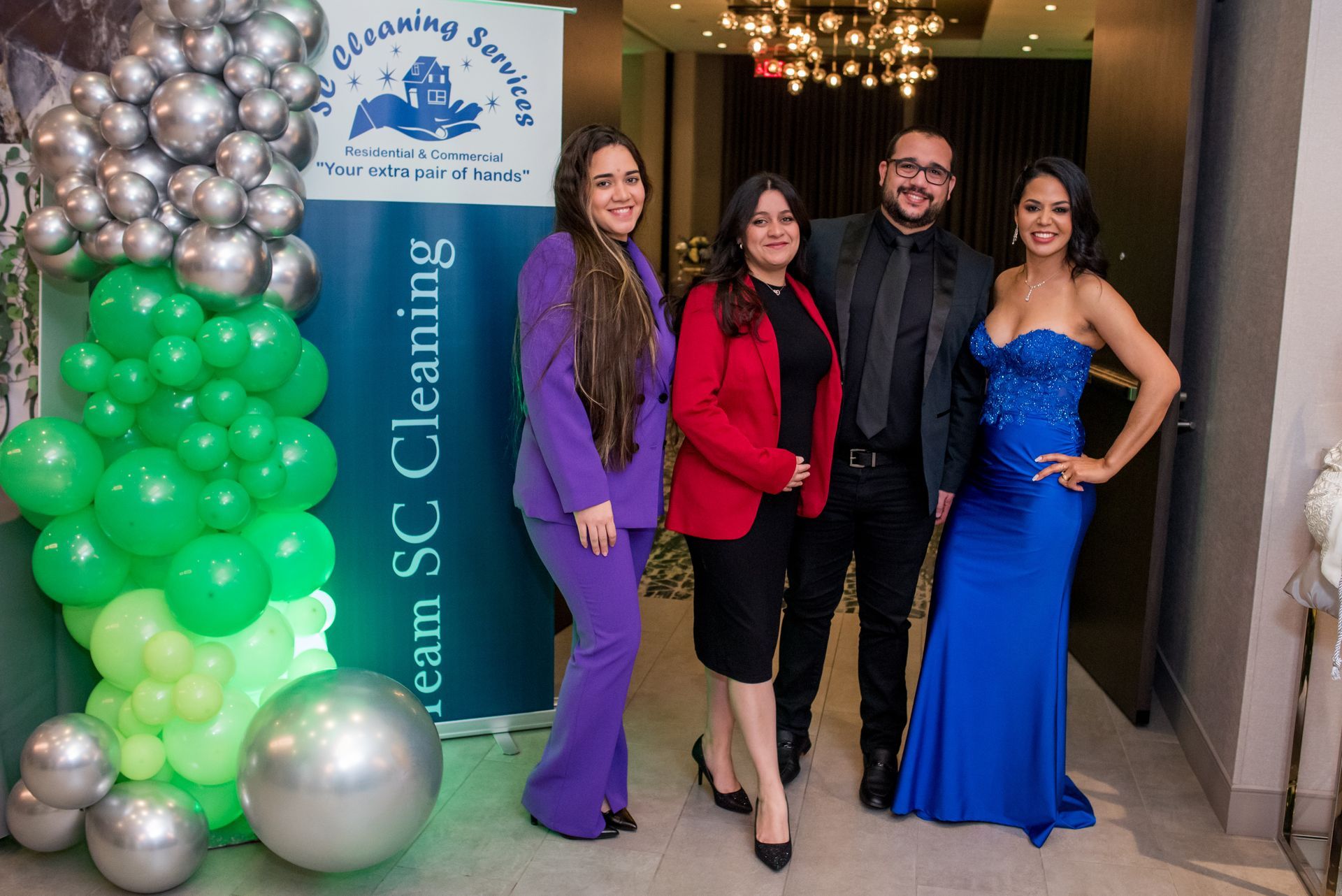 Four people posing; woman in purple suit, red jacket, black suit, and blue gown by balloon arch.