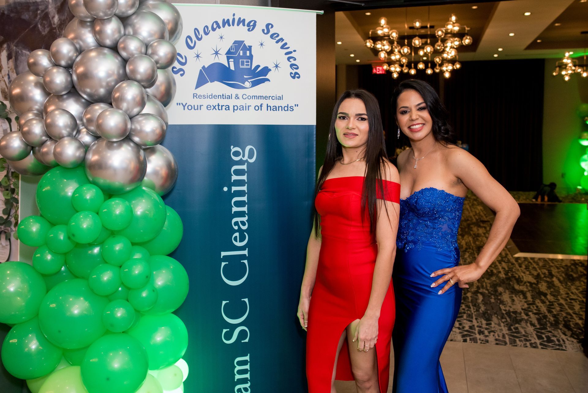 Two women in formal dresses pose by a banner with balloons. One in red, one in blue, smiling.