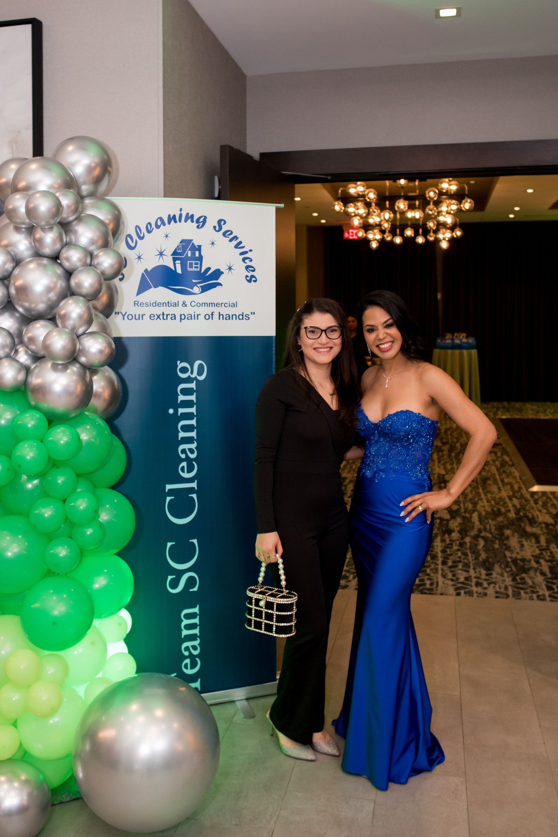 Two women pose by a banner for a cleaning service, one in a blue gown and the other in a suit.