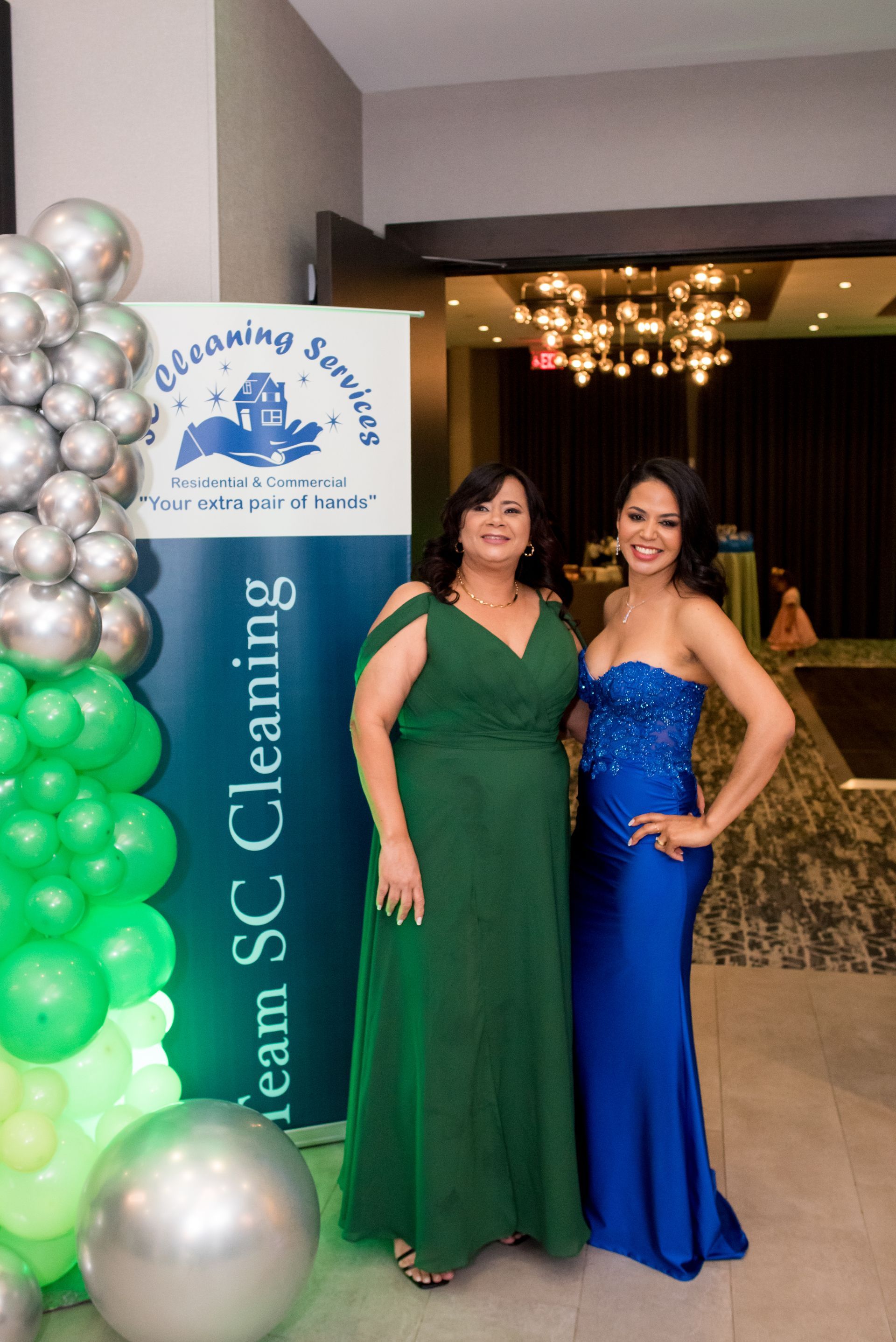 Two women in gowns pose by a banner and balloons at an event. One in emerald green, the other in blue.