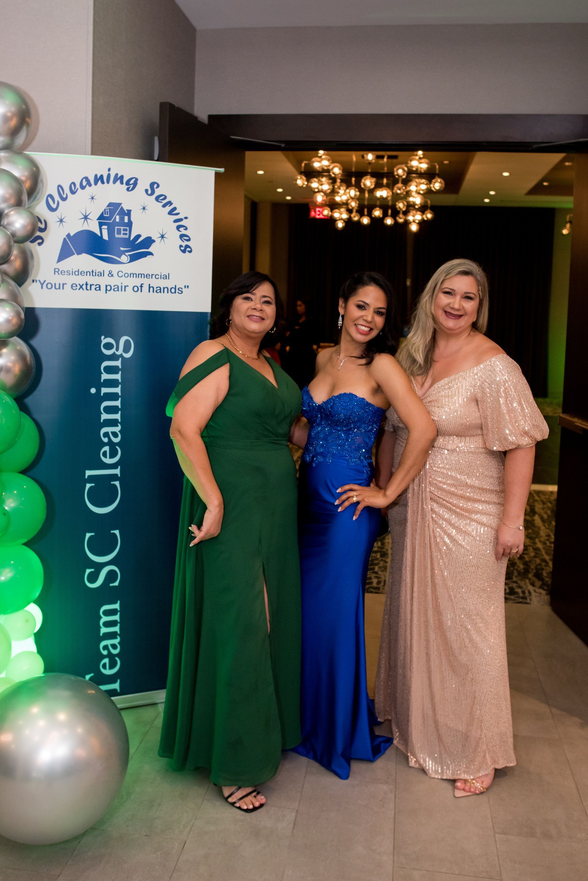 Three women in gowns pose by a banner at an event. One in green, one in blue, and one in pink/gold.