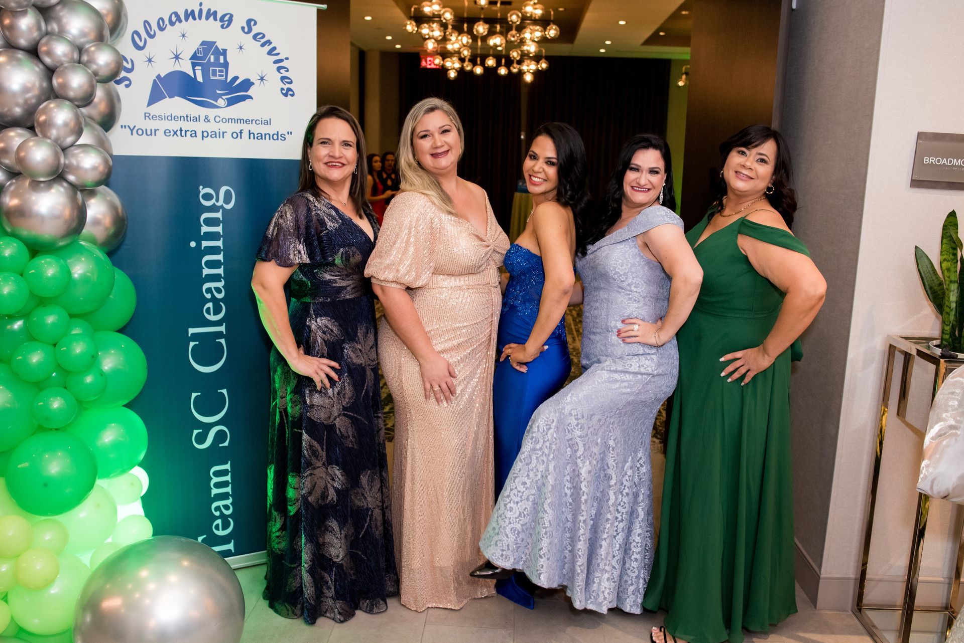 Five women in formal gowns pose at an event. A green and silver balloon arch is in the background.
