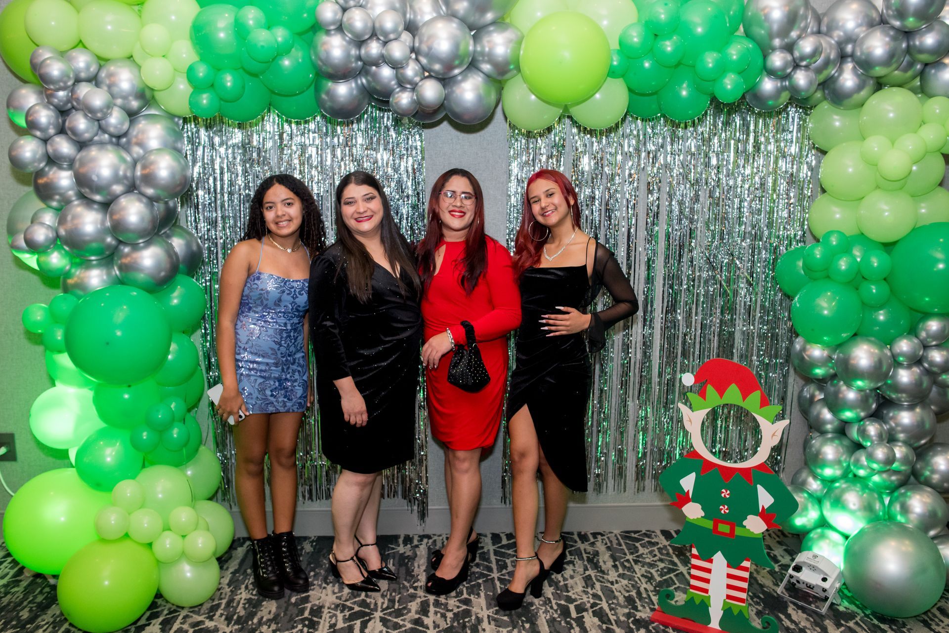 Four women pose in front of a balloon arch and foil fringe backdrop. Green, silver balloons. Holiday cutout.