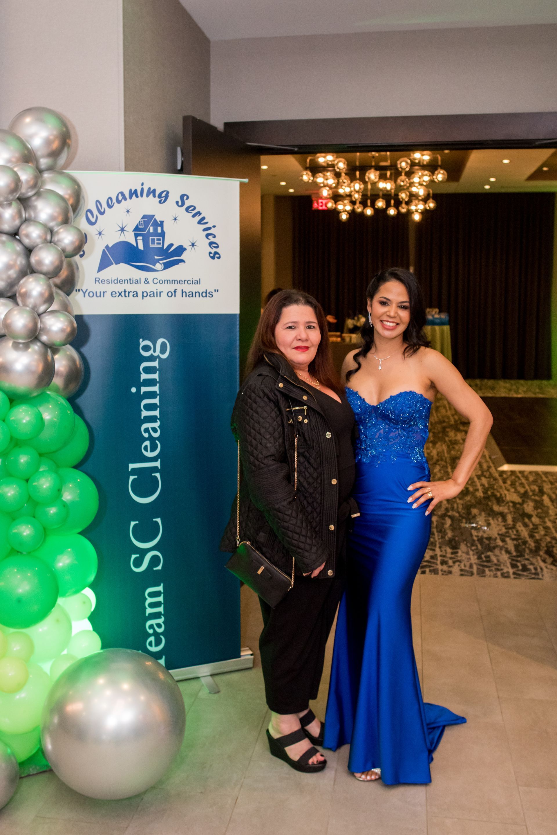 Two women pose in front of a banner for a cleaning service at an event with balloons.